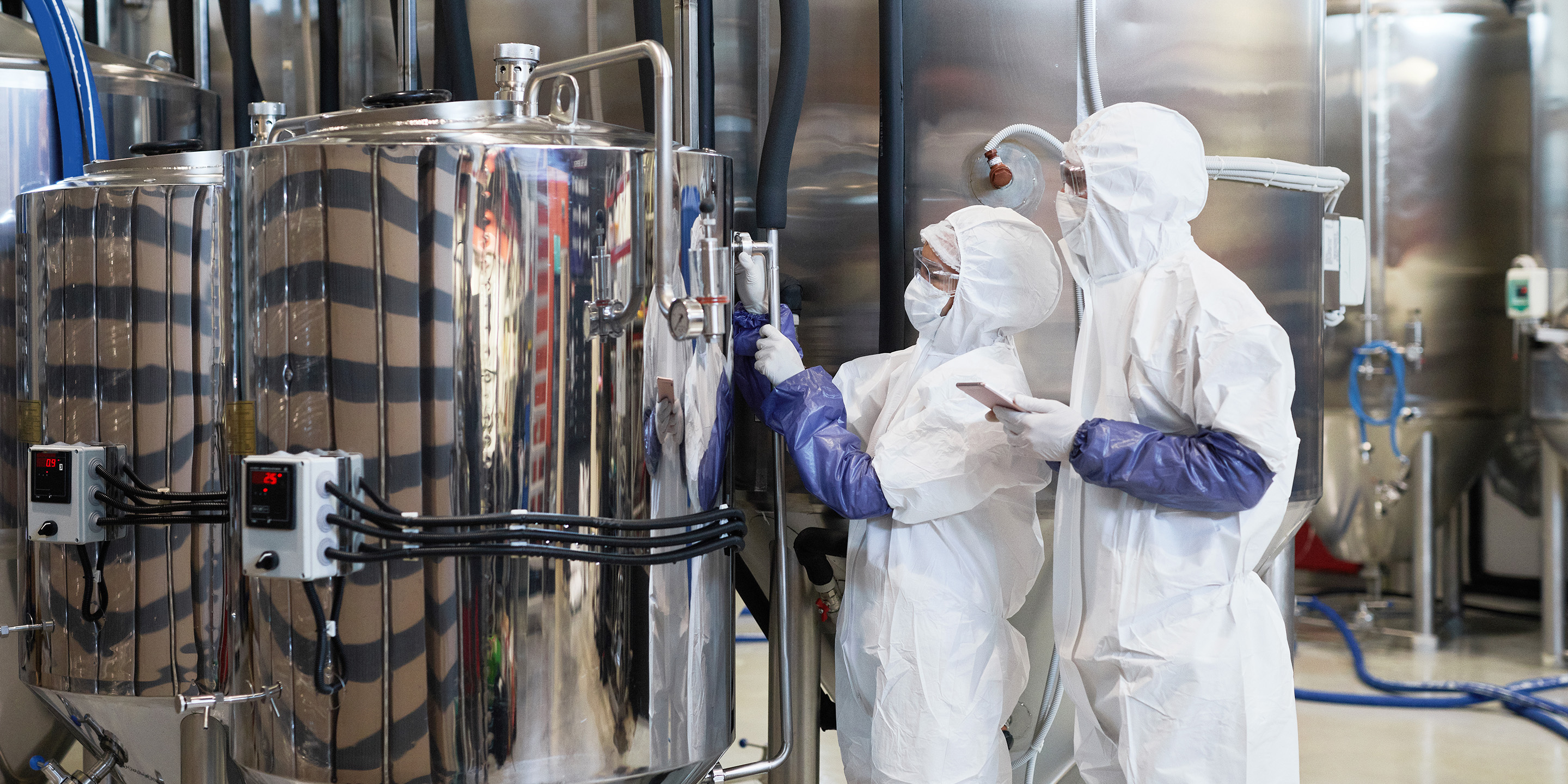 Two laboratory technicians in white protective suits are inspecting a large metallic tank. One uses a tool, while the other holds a smartphone. The background features industrial machinery and piping.