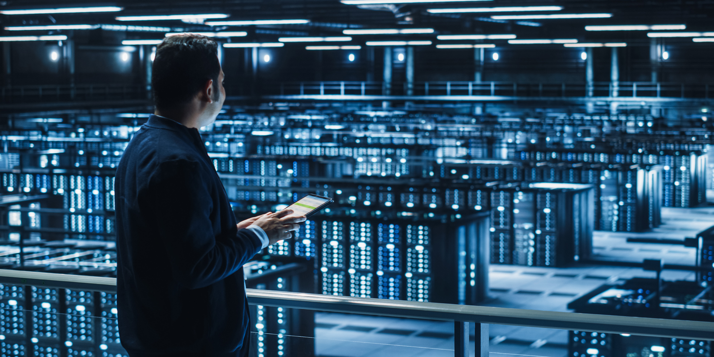 A person with a tablet stands on a balcony above a brightly lit data center. Rows of active server racks with blinking lights stretch below, showcasing a high-tech infrastructure environment.