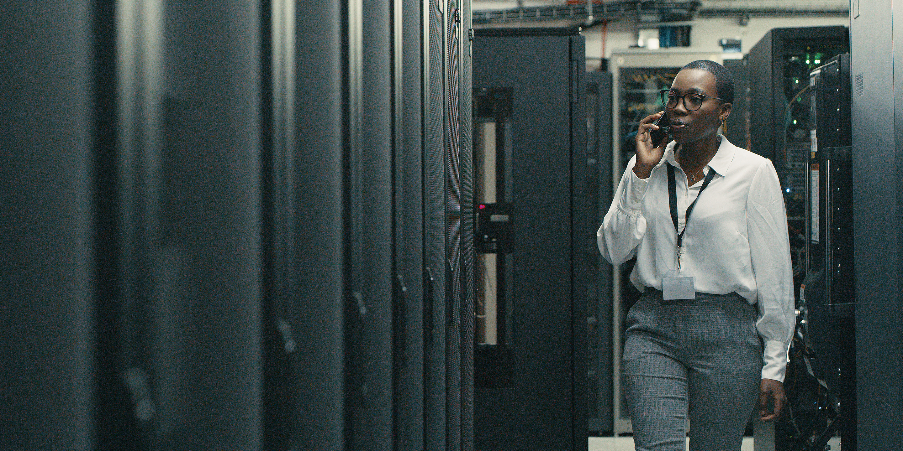 A person in business attire walks through a data center, engaged in a phone conversation, surrounded by rows of server racks.
