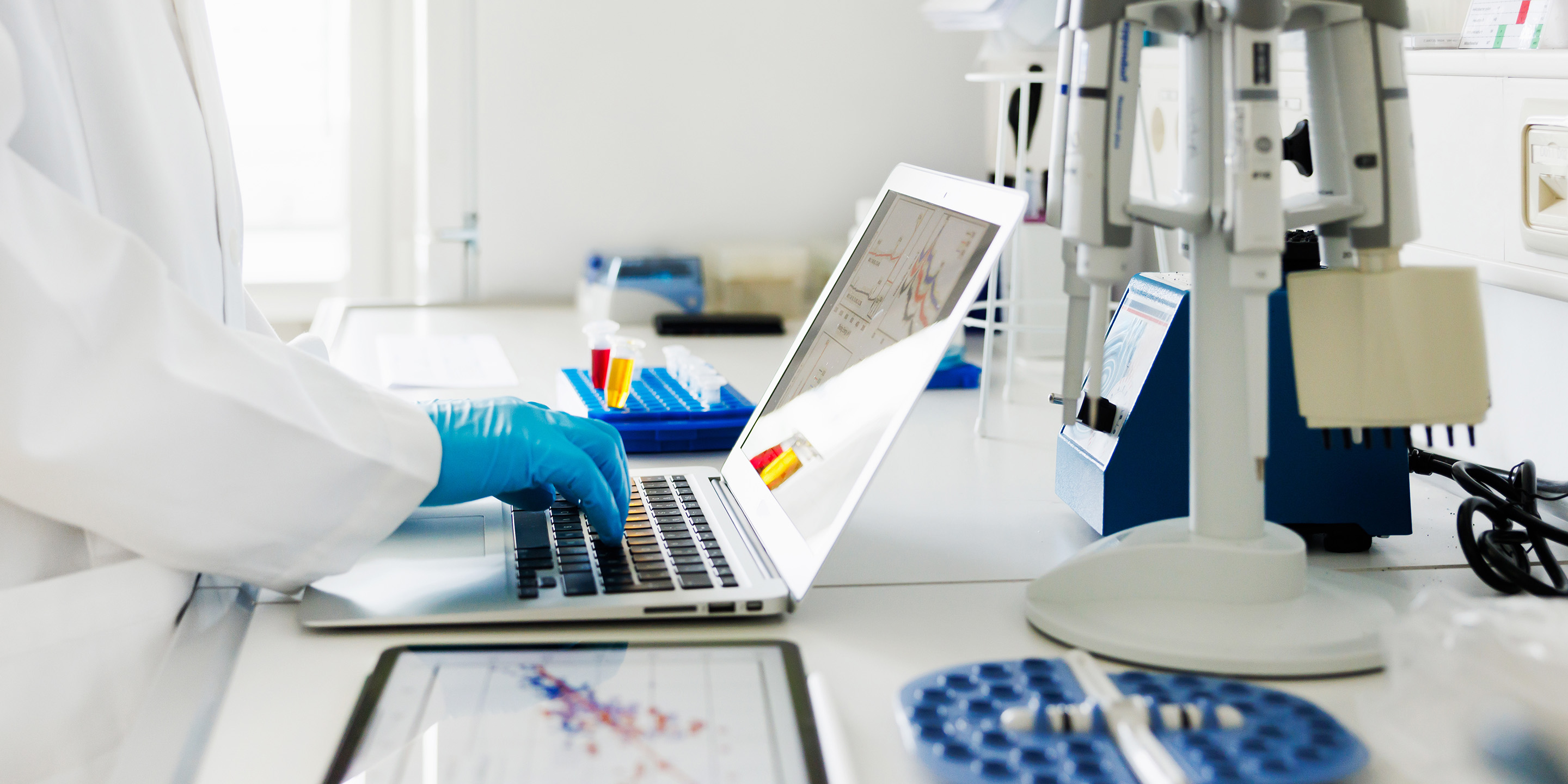Scientist wearing blue gloves working on a laptop in a laboratory, surrounded by test tubes, a tablet displaying data charts, and other scientific instruments.