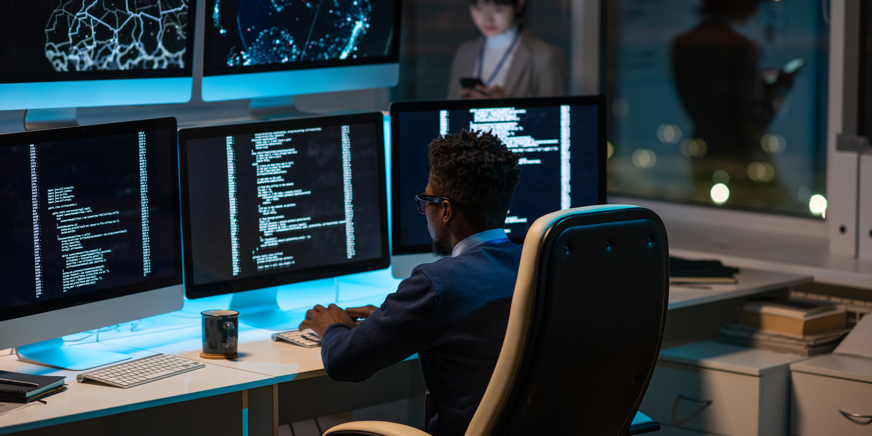 A software developer working at a desk with multiple monitors displaying code and data visualizations in a modern office setting. Another person is seen in the background using a smartphone.