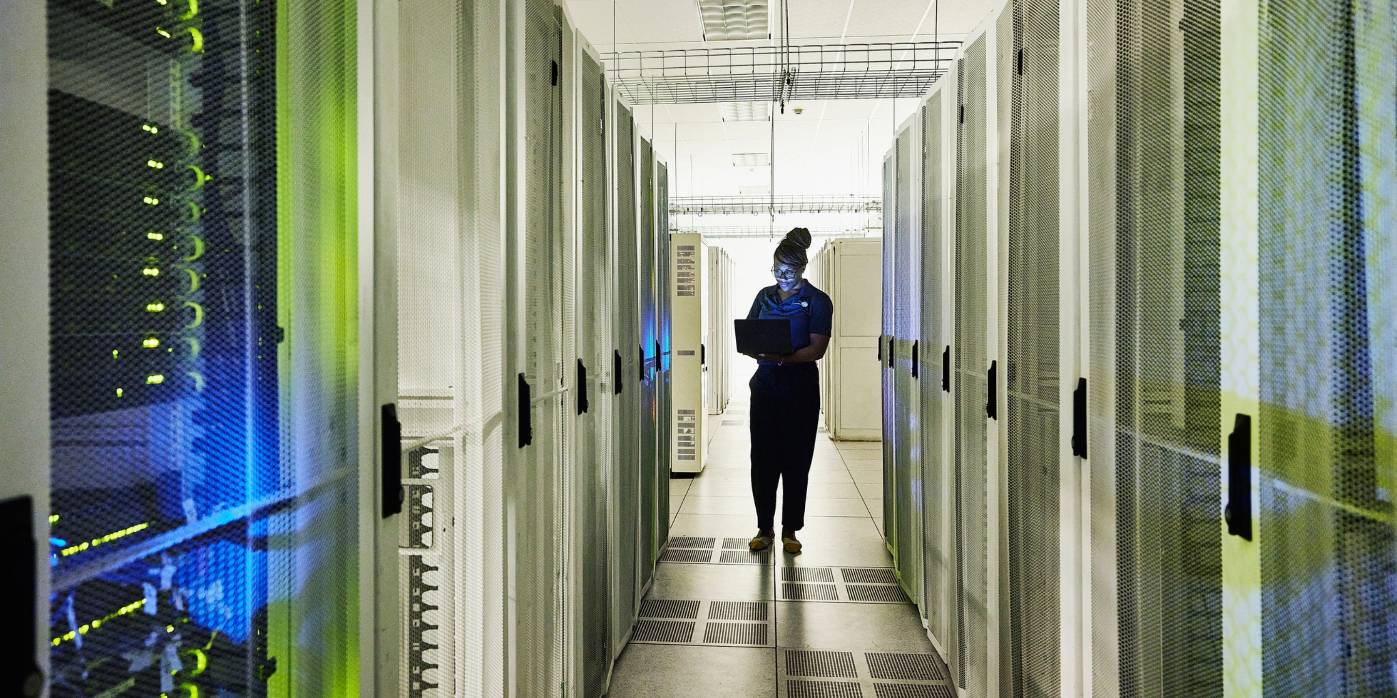 A person stands in a brightly lit server room, holding a laptop amid rows of tall server racks. The environment features visible cabling and tiled flooring, highlighting a high-tech data infrastructure setting.