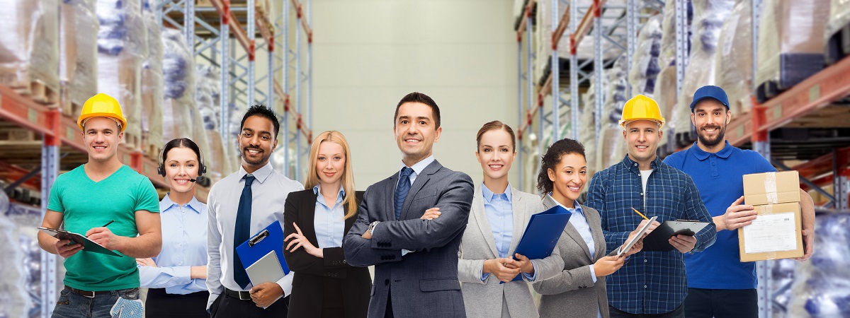 A team of nine professionals in a warehouse with tall shelves and stacked pallets. Some hold clipboards and tablets, highlighting logistics oversight.
